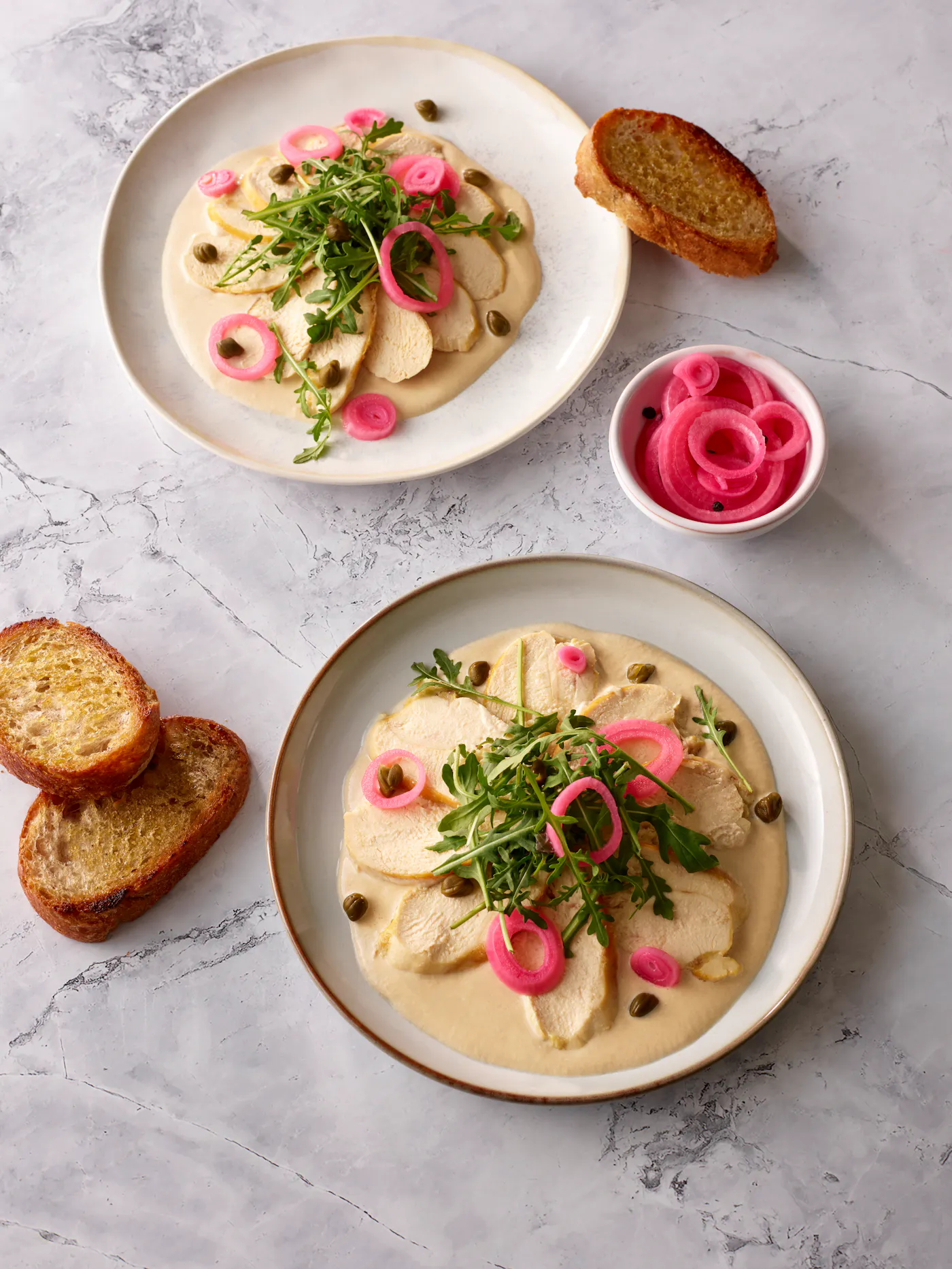Pollo Tonnato serviert mit Rucola und Pickled Onions auf hellem Gedeck mit geröstetem Brot