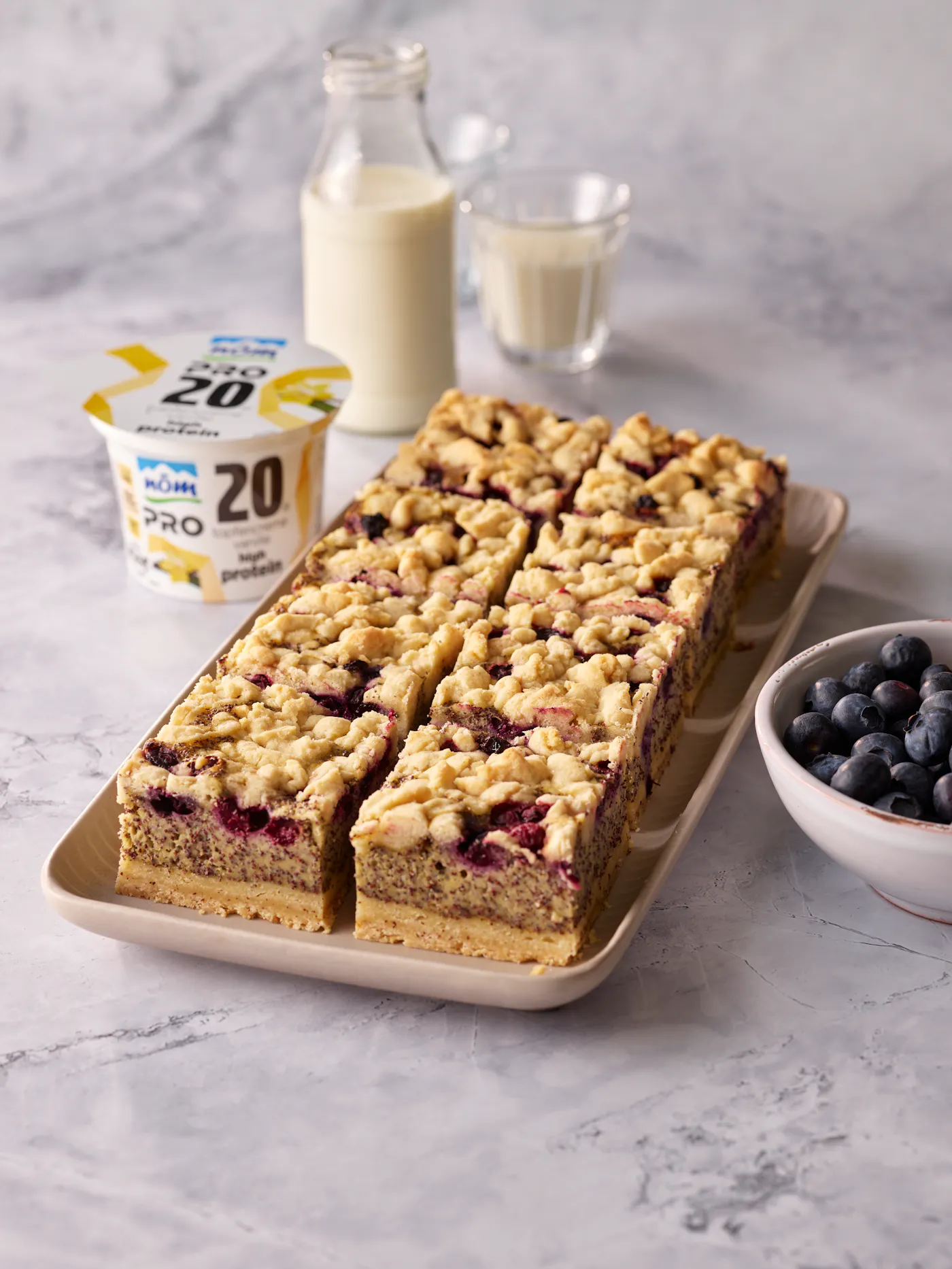 Auf dem Bild ist ein Mohn-Streuselkuchen zu sehen. Direkt daneben steht eine Schüssel voller Heidelbeeren, im Hintergrund Milch und eine NÖM PRO Topfencreme Vanille,