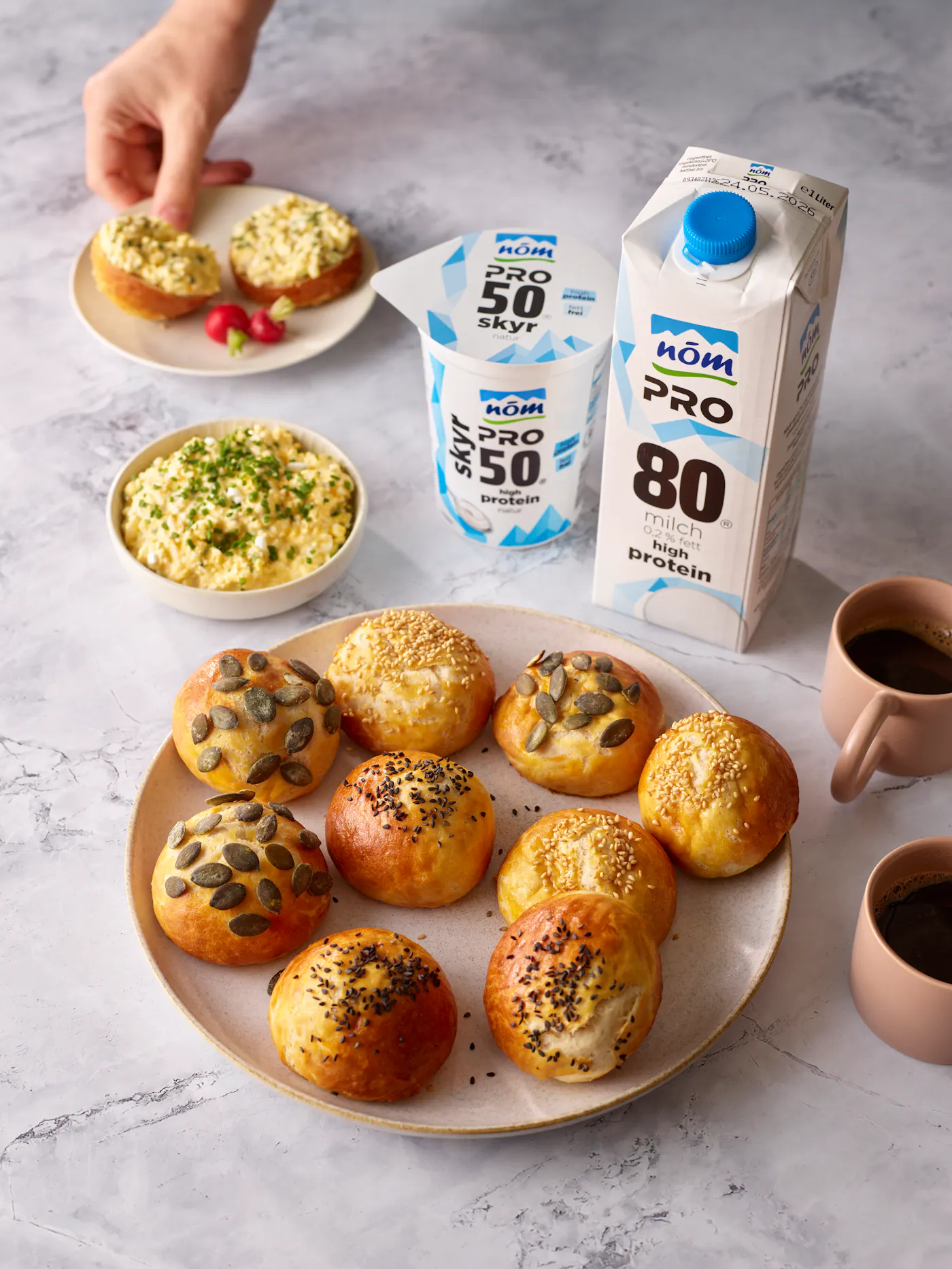 Laugenweckerl mit NÖM PRO Skyr im Teig umgeben von einem Ei-Aufstrich, NÖM PRO Produkten und schwarzem Kaffee