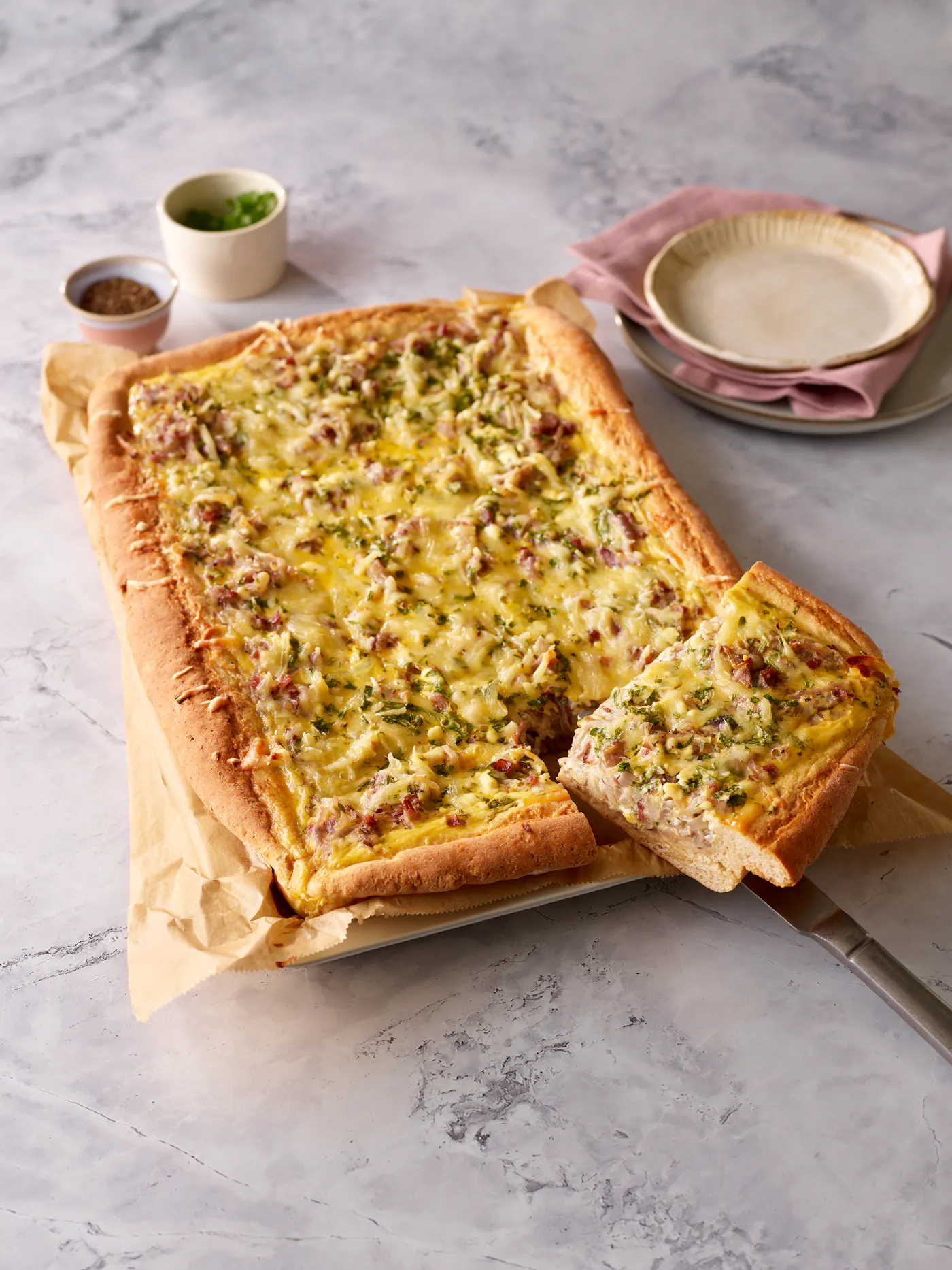 Angeschnittener, herzhafter Zwiebelkuchen auf einer marmorierten Platte mit Tassen und Tellern im Hintergrund