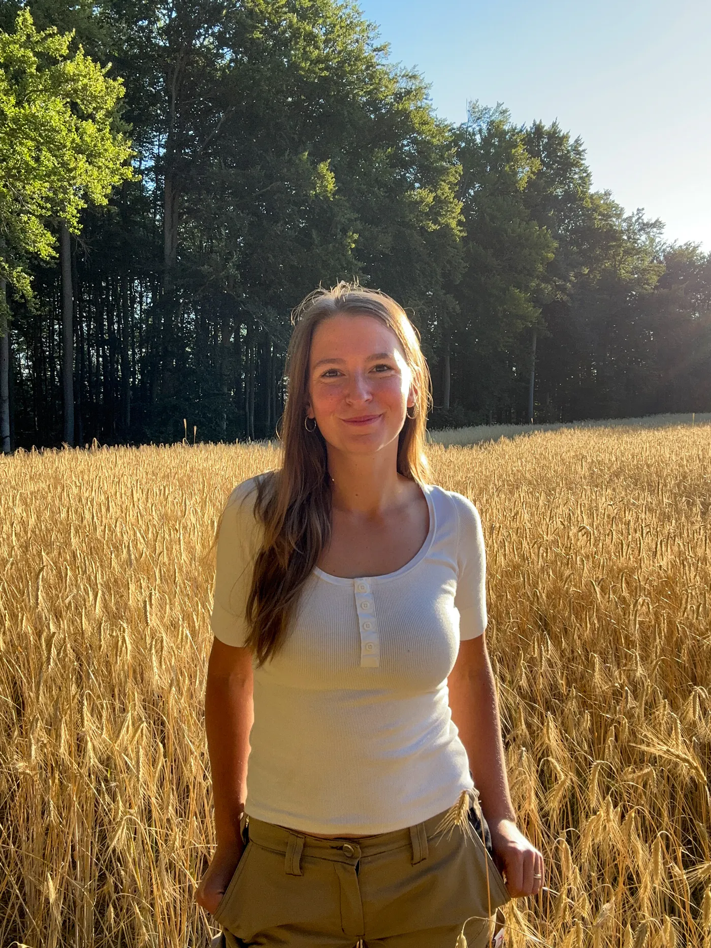 Bäuerin Johanna in einem Weizenfeld Die Bäuerin Johanna trägt ein weißes T-Shirt und steht in einem Weizenfeld vor grünen Bäumen und blauem Himmel