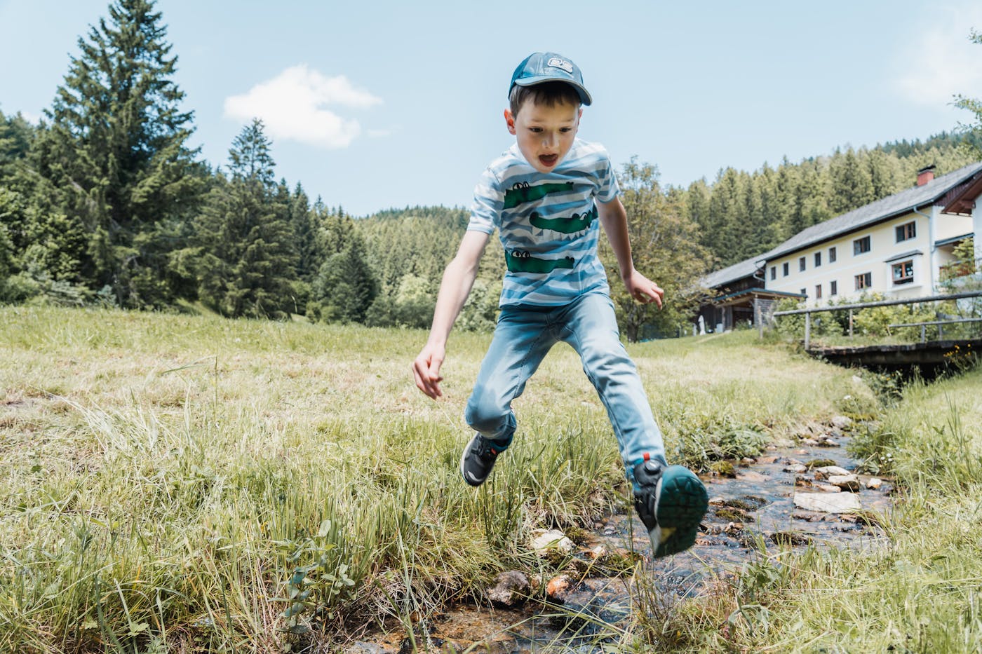 Fotocredit Gerald Demolsky 3 Kind beim Sprung über Bach mit Haus im im Hintergrund