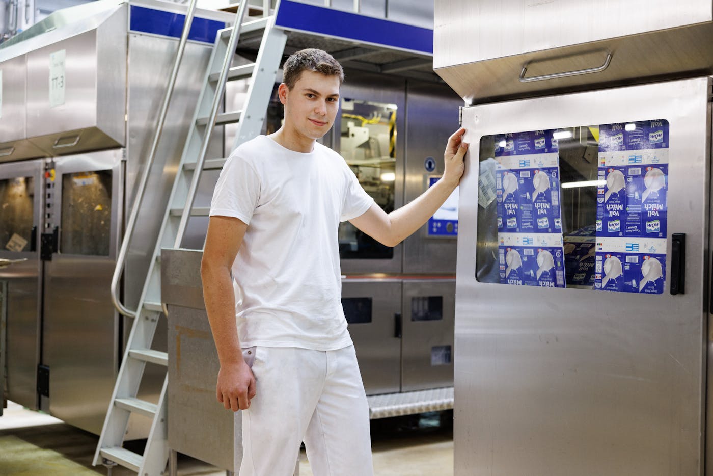 junger Mitarbeiter der NÖM in der Produktionshalle neben einer Maschine mit Milchpackungen junger Mitarbeiter der NÖM in der Produktionshalle neben einer Maschine mit Milchpackungen