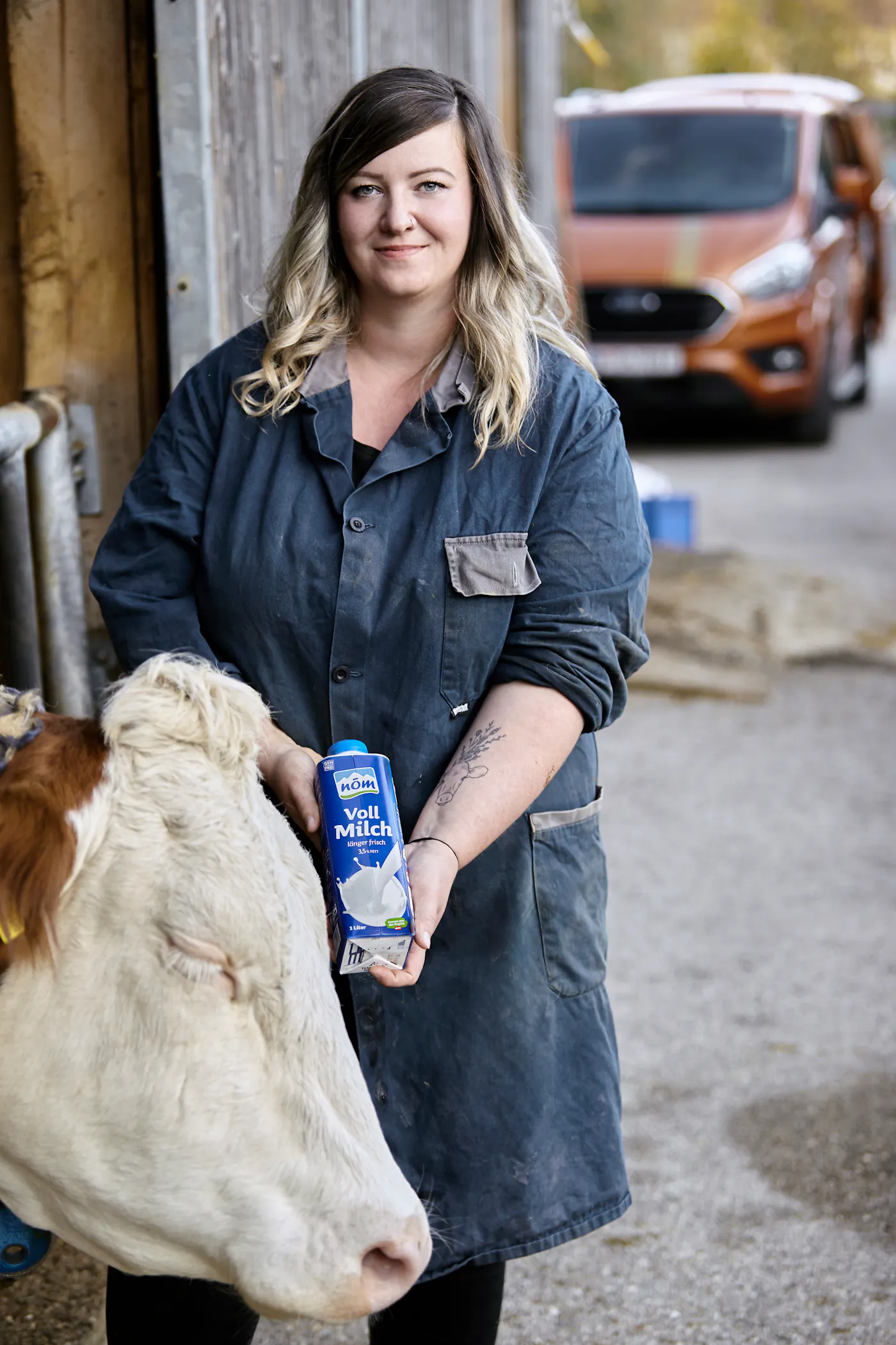 NÖM Bäuerin Corinna Büchner mit einer NÖM Vollmilch und einer Kuh NÖM Bäuerin Corinna Büchner steht in Arbeitskleidung vor dem Stall mit einer Kuh und einem Liter NÖM Vollmilch