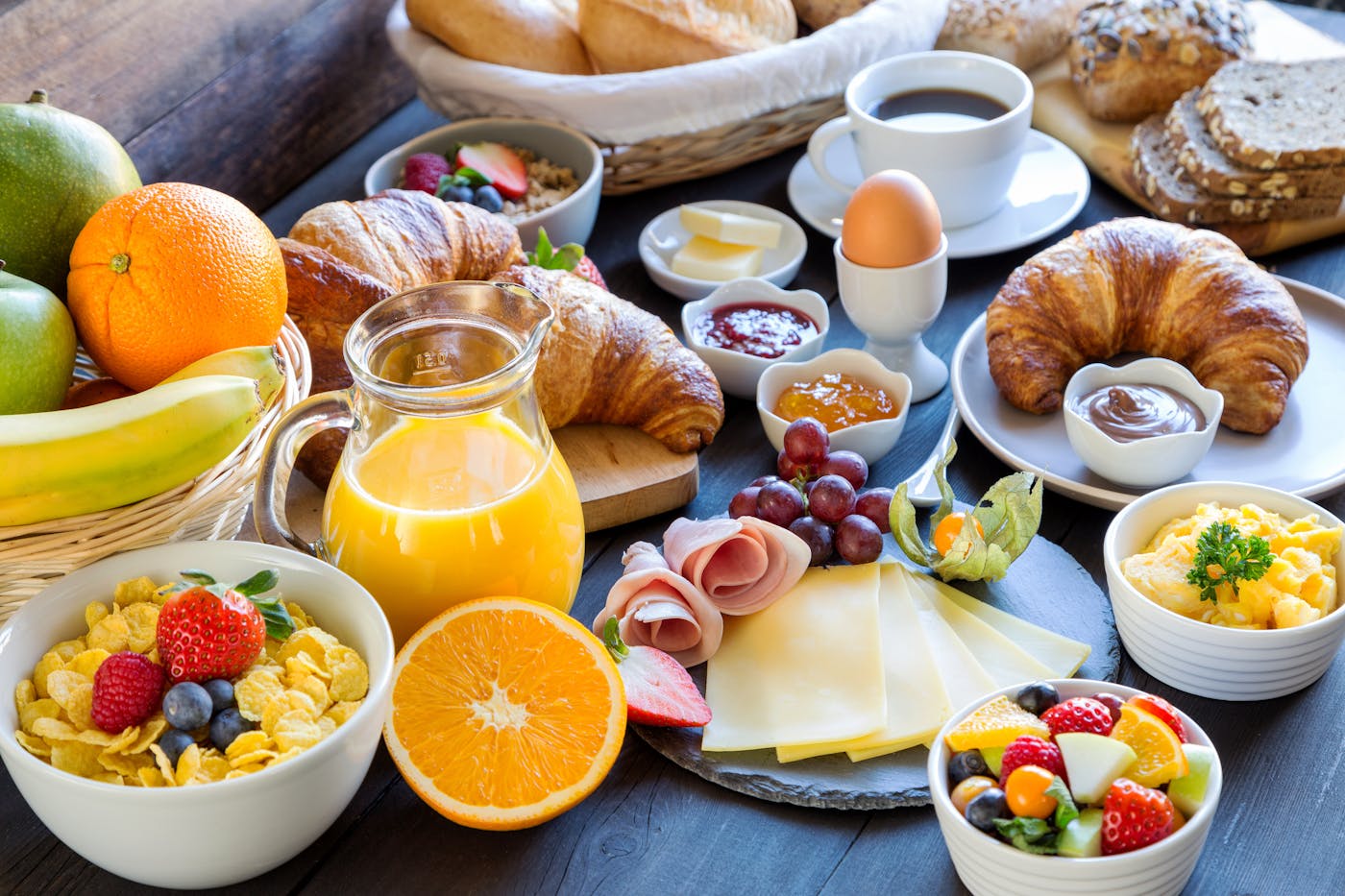 Reich gedeckter Frühstückstisch mit Obst, Müsli, Orangensaft, Croissants, Käse und Eiern.