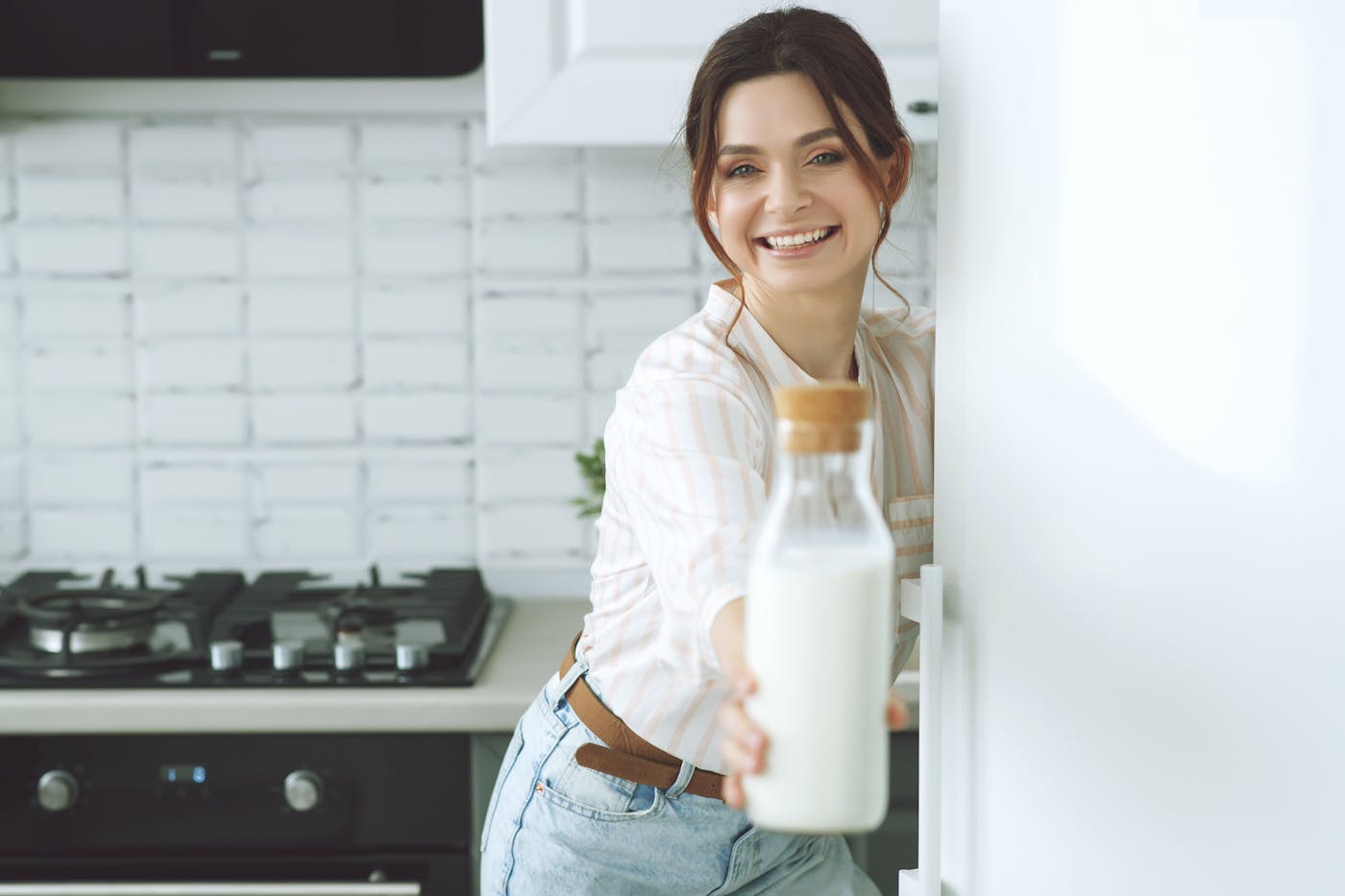 Braunhaarige Frau steht in der Küche und hält eine Glasflasche mit Milch in die Kamera