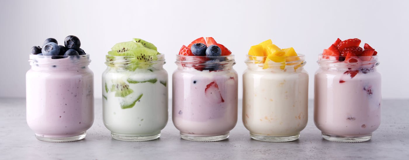 Reihe von fünf Joghurts in Gläsern, garniert mit verschiedenen frischen Früchten wie Heidelbeeren, Kiwis, Erdbeeren und Mango