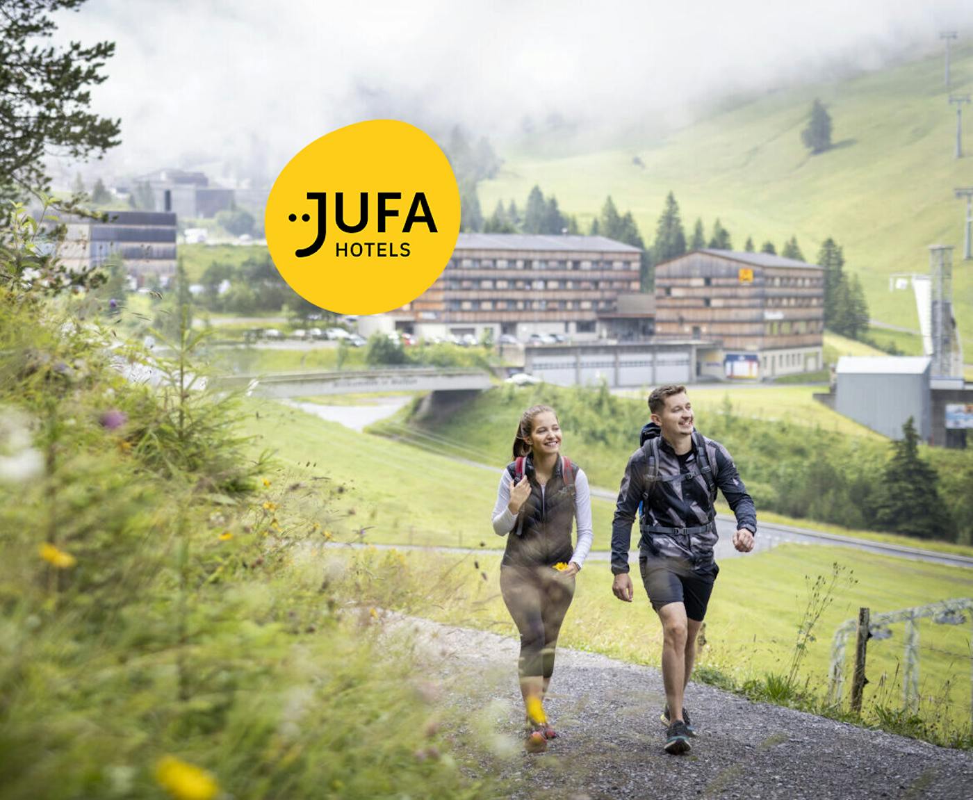 Paar wandert in Wanderausrüstung auf einem Forstweg im Hintergrund das Jufa Hotel Malbun