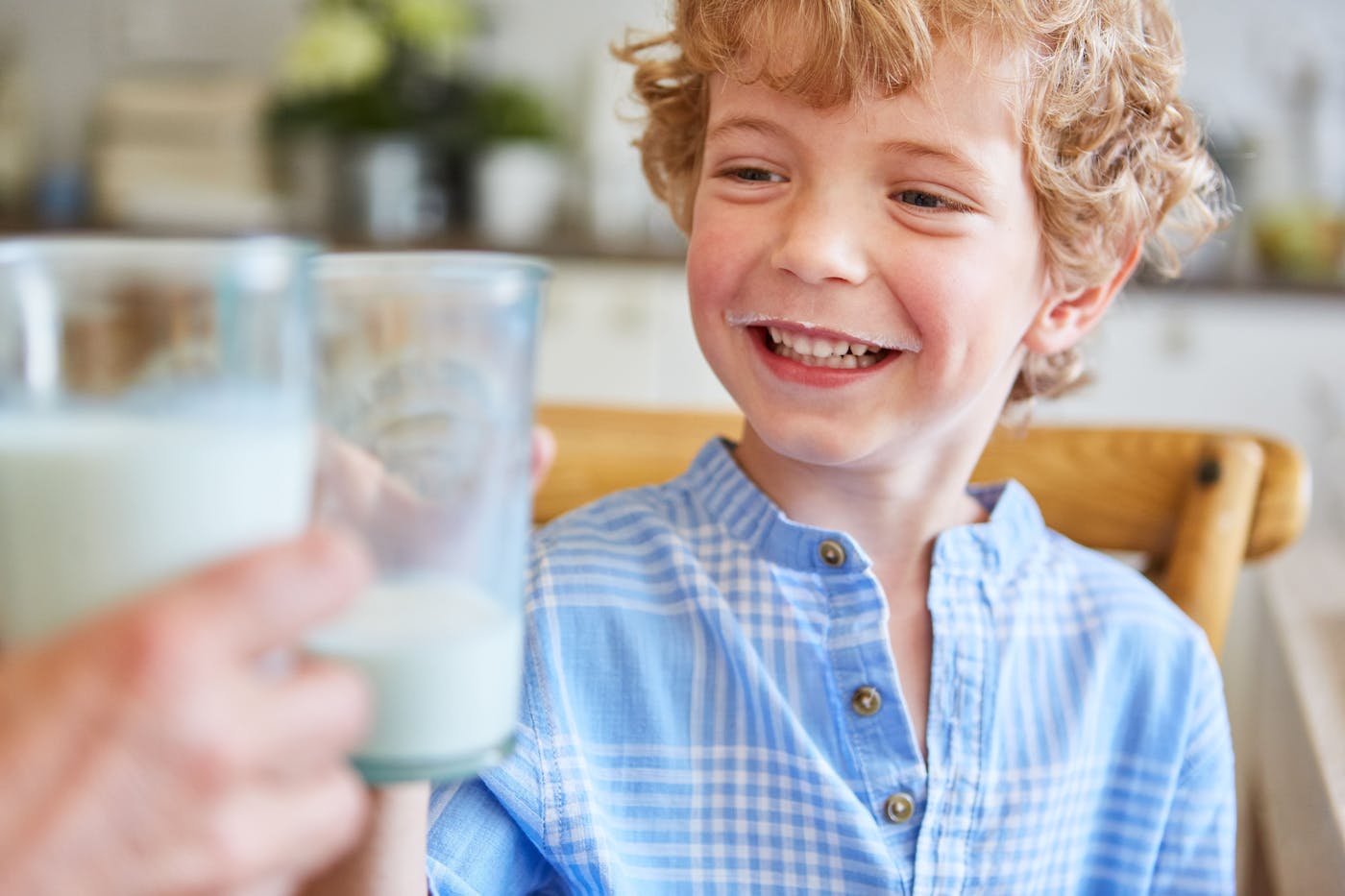 Lachender Junge in blauem Hemd und Milchbart prostet jemandem mit einem Glas Milch zu