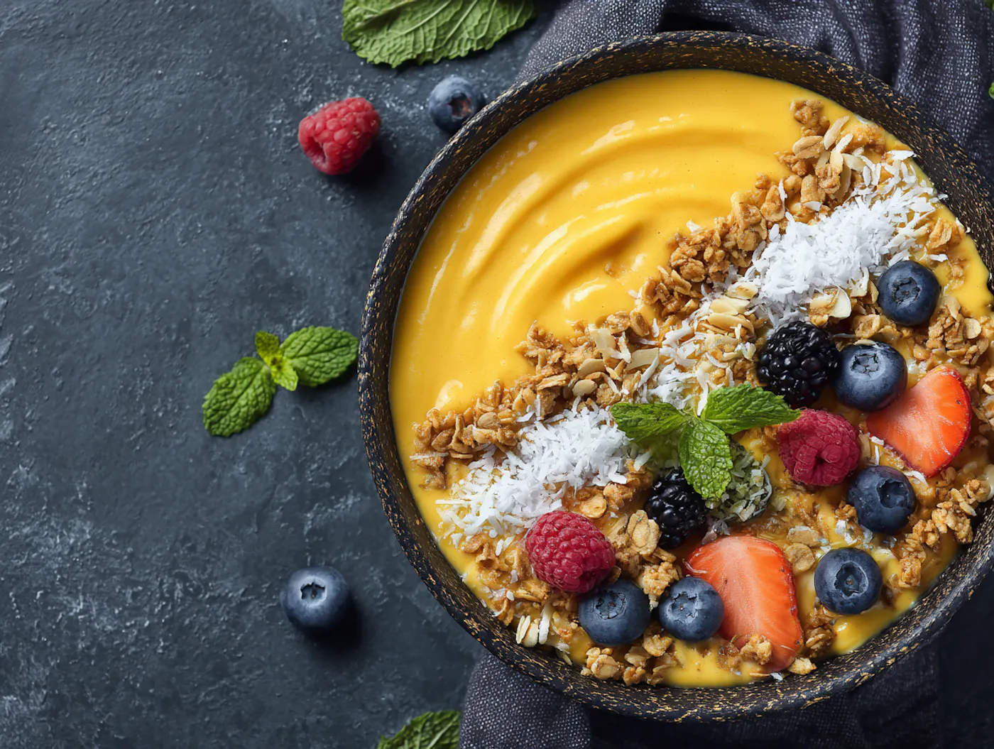 Eine Mango Smoothie Bowl in einer dunklen Schüssel getoppt mit Kokosflocken, Granola, frischen Beeren und etwas Minze