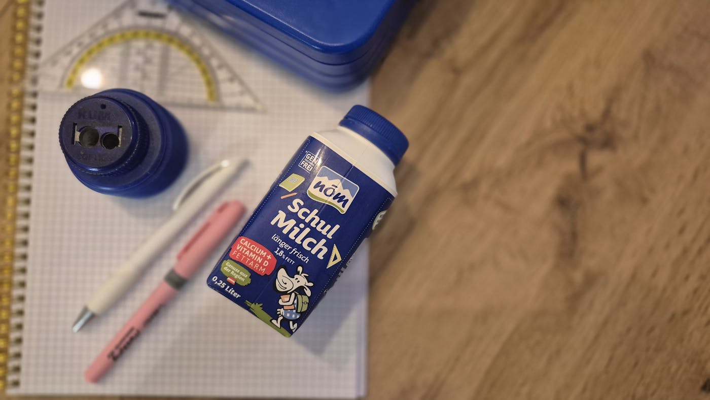 Schulmaterialien mit NÖM Schulmilch-Packung auf Holzfläche: Trinkflasche, Stifte, Geodreieck, Brotdose und kariertes Papier.