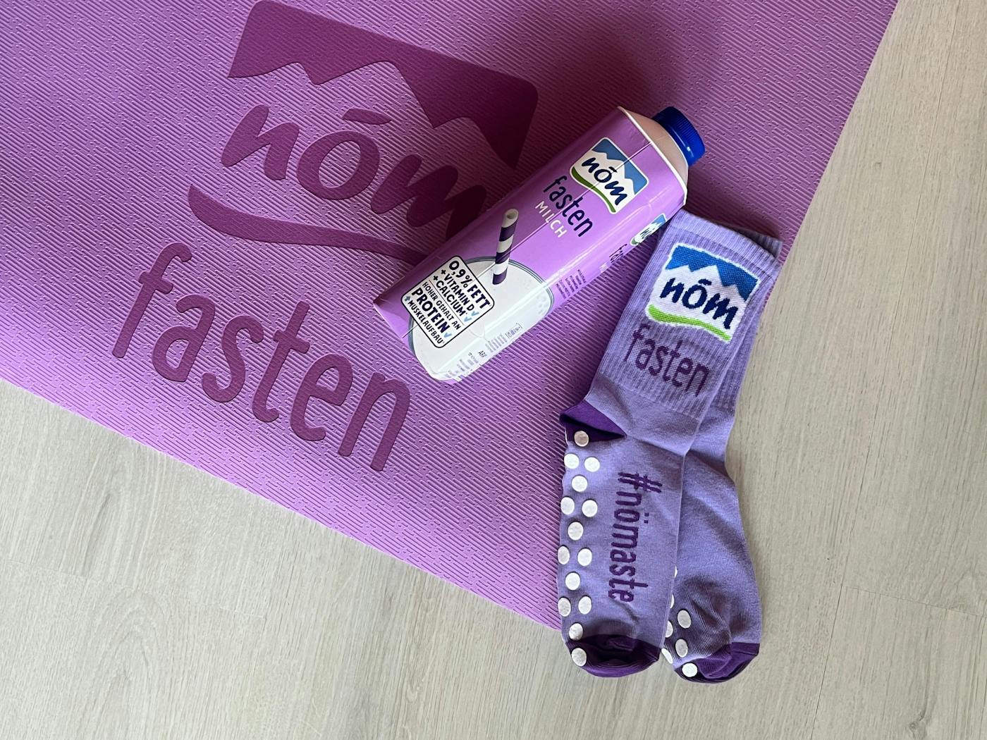 Lila Yogamatte mit „NÖM fasten“-Aufdruck, darauf lila NÖM fasten-Drink und gepunktete Socken mit „NÖM fasten“-Schriftzug.